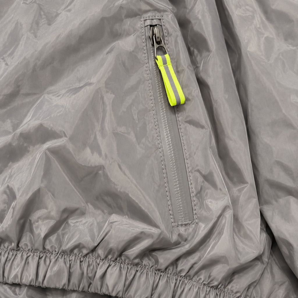 Close-up of a grey windbreaker pocket zipper, neon yellow-green pull and gathered hem.