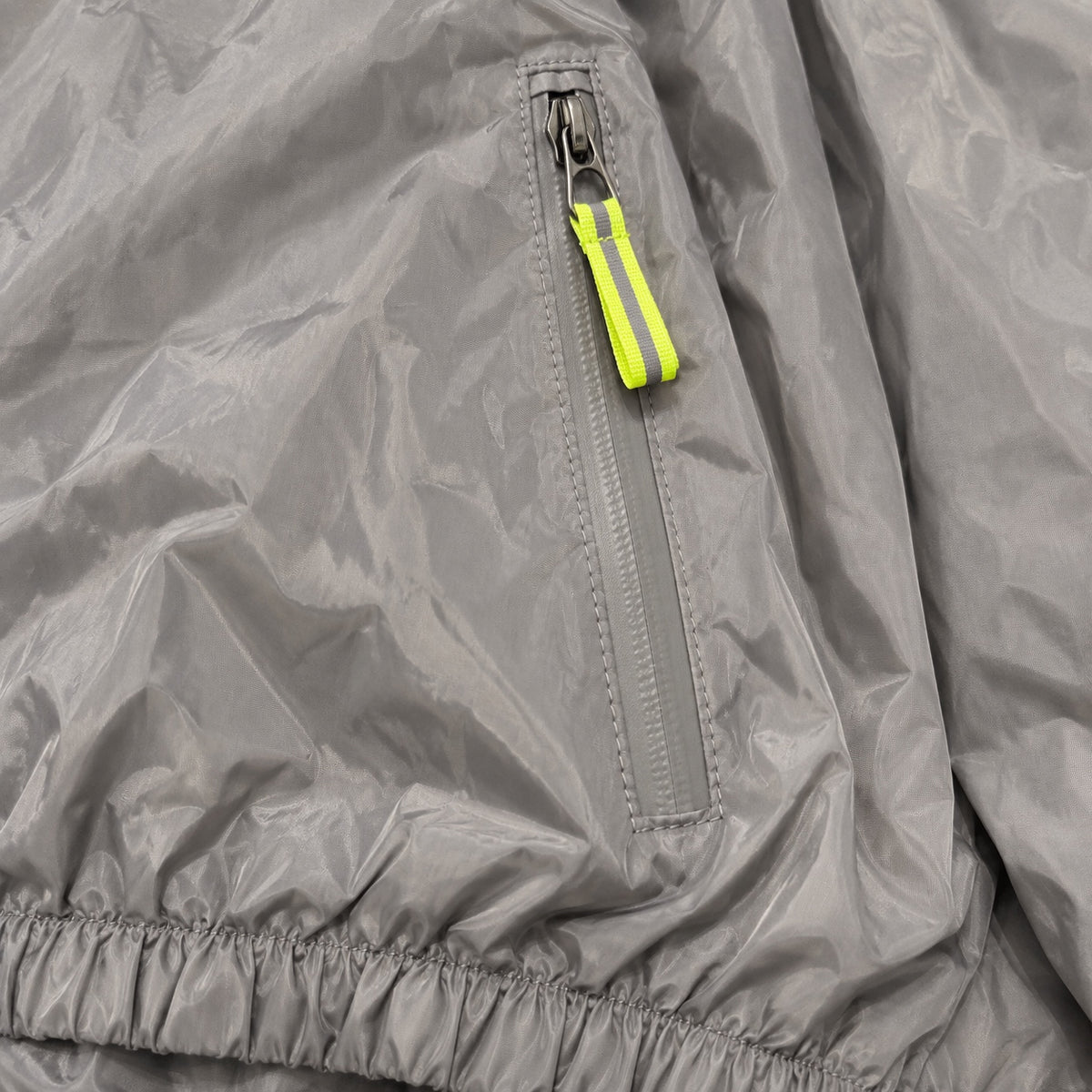 Close-up of a grey windbreaker pocket zipper, neon yellow-green pull and gathered hem.